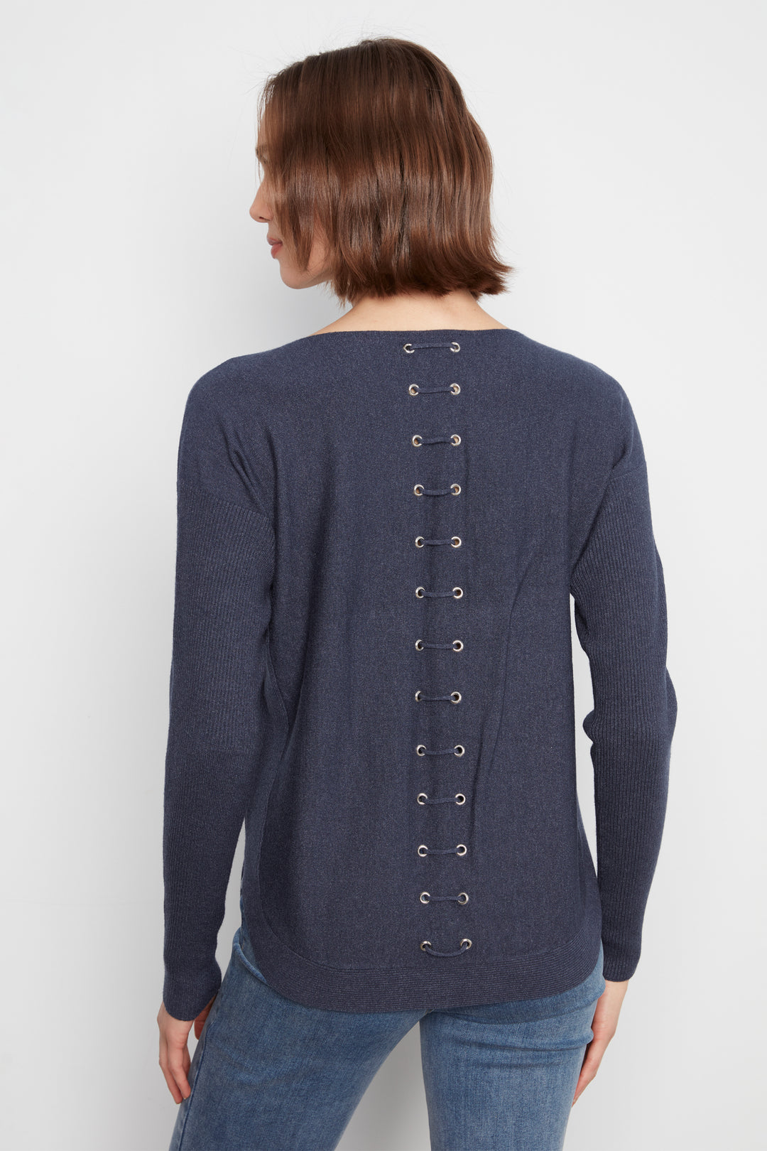 A woman shown from the back wearing a navy blue long-sleeve knit top with a vertical lace-up detail down the back, paired with blue jeans against a plain light gray background.