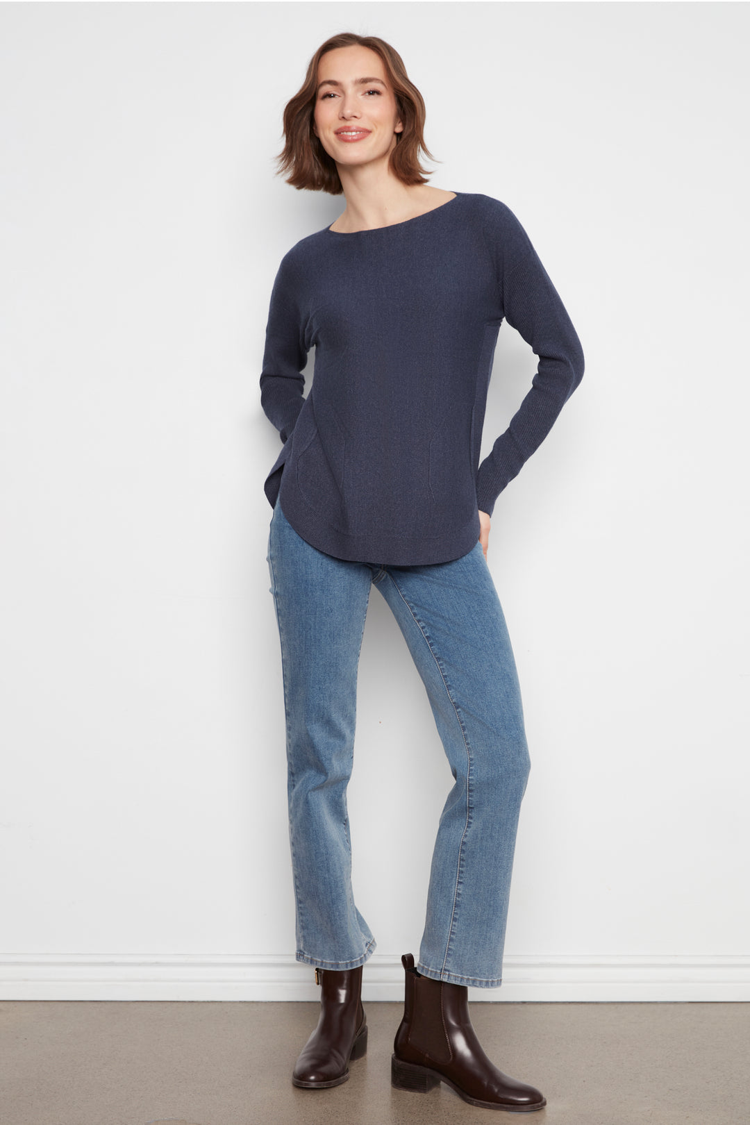 A woman wearing a dark navy blue long-sleeve knit top with a wide boat neckline, paired with blue jeans, against a plain light gray background.
