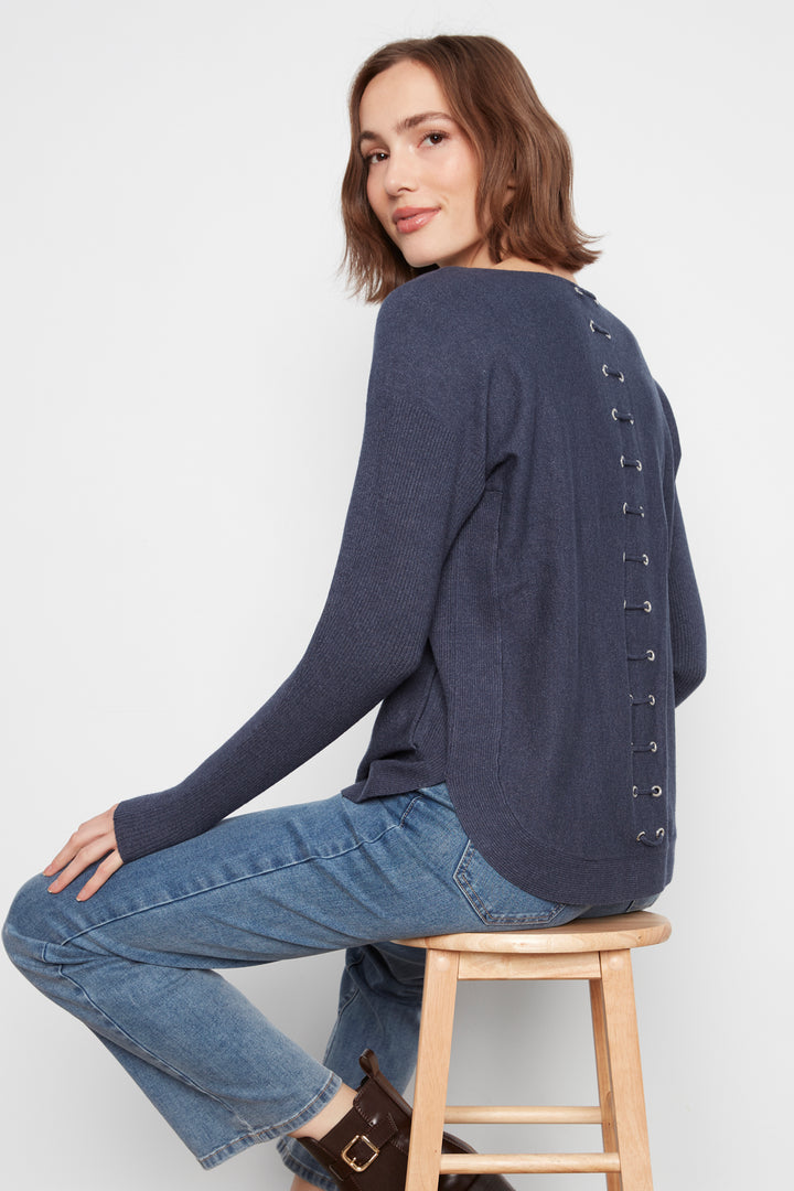 A woman wearing a navy blue knit sweater with lace-up detailing down the back, paired with blue jeans, sitting on a wooden stool against a light gray background.