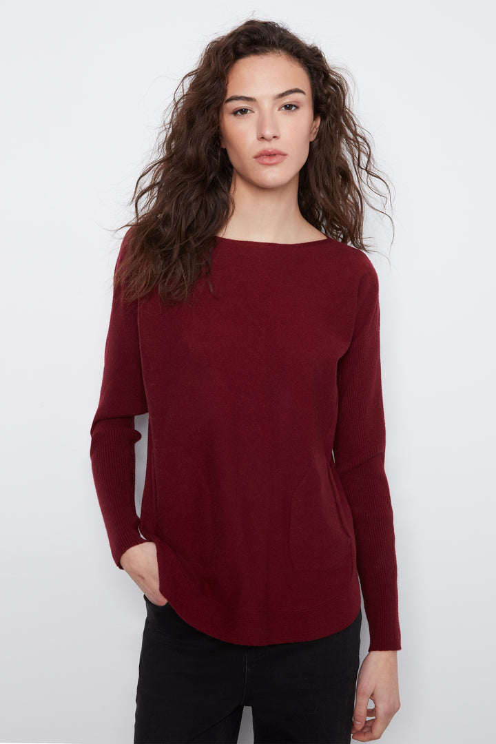 A woman with long, wavy brown hair wearing a long-sleeve boat-neck knit sweater in deep burgundy, paired with black pants, against a plain light gray studio background.
