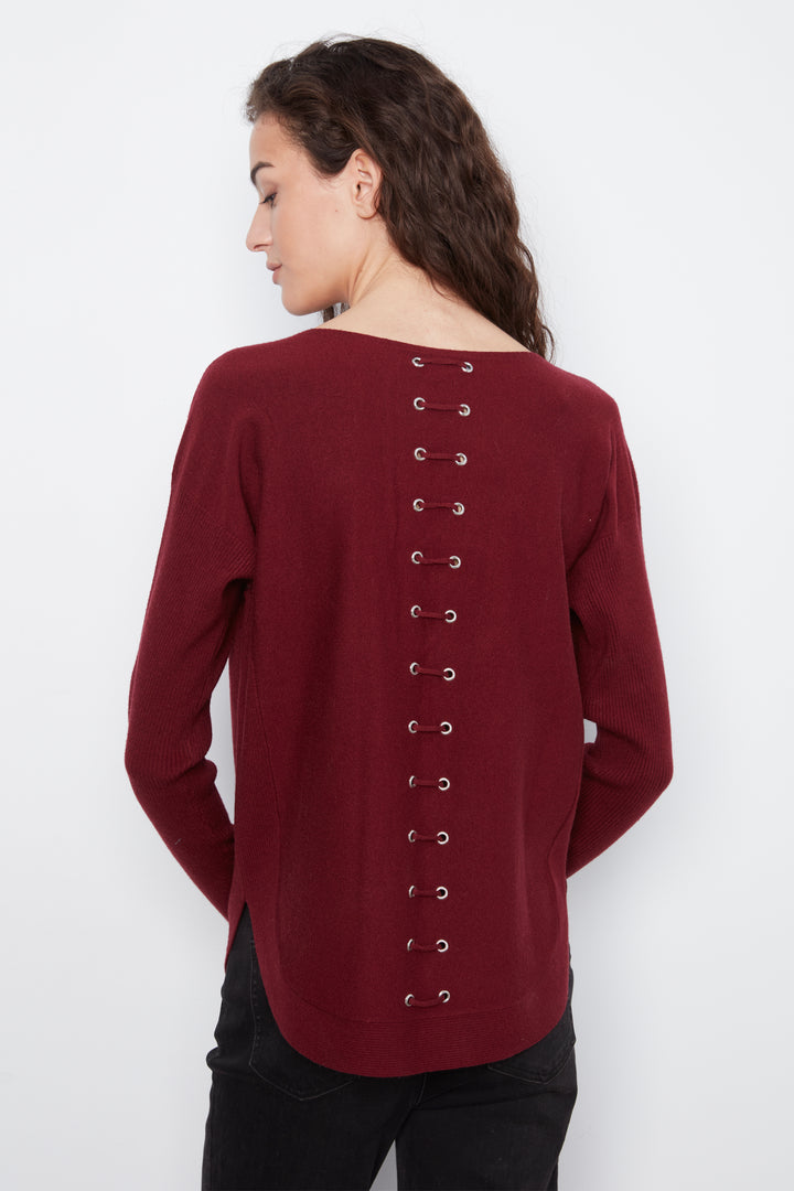 A woman shown from the back wearing a long-sleeve maroon knit top with a vertical row of eyelet lace-up details down the center, paired with black pants, against a plain white background.