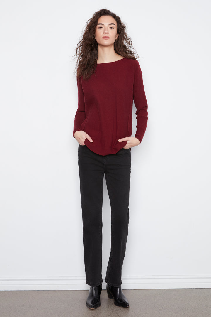 A woman stands against a white wall wearing a long-sleeve boat-neck knit sweater in deep burgundy, paired with black pants and black ankle boots.