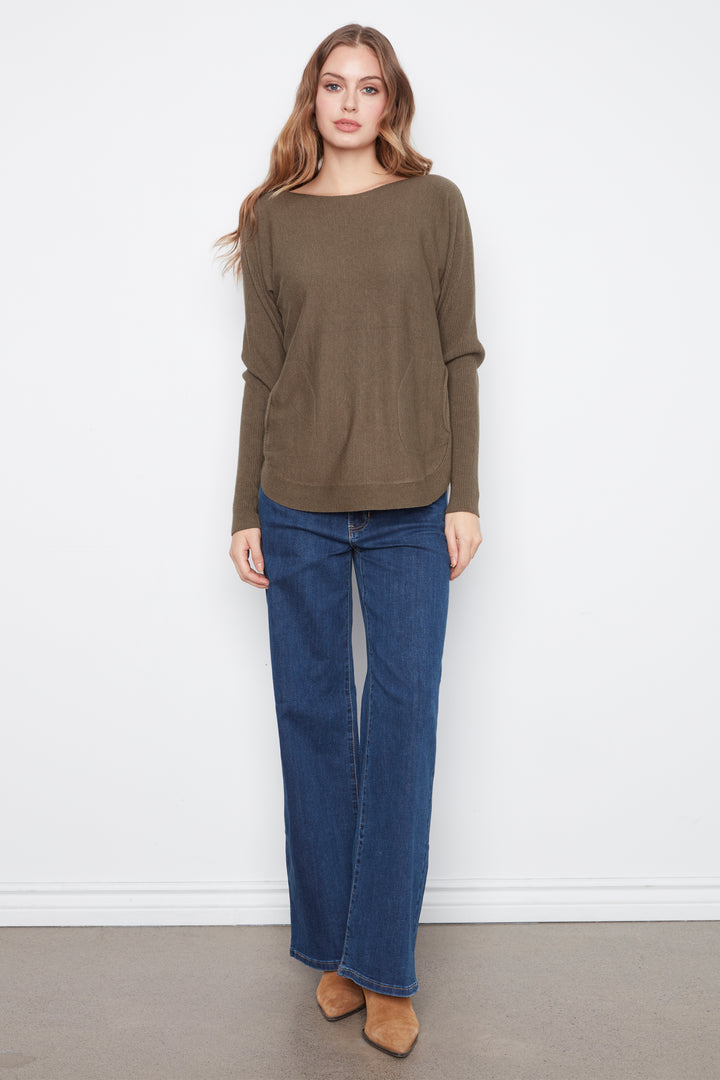 A woman modeling a long-sleeve olive green knit top with wide boat neck and straight cuffs, paired with blue jeans and tan suede boots, standing against a white wall.