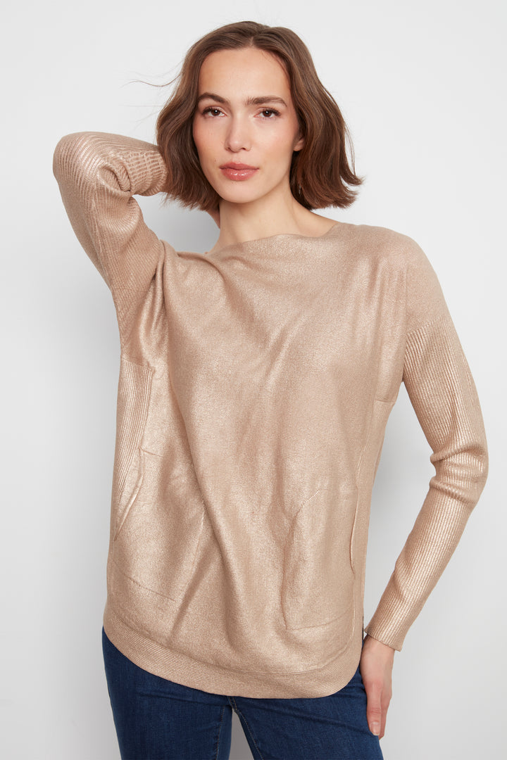 A woman wearing a metallic champagne-beige long-sleeve knit top with a draped neckline, paired with dark blue jeans, photographed against a light gray studio background.