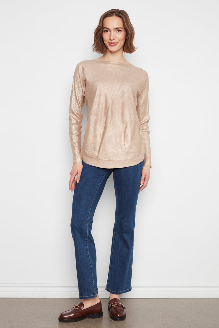A woman modeling a light beige long-sleeve knit sweater, paired with dark blue jeans and brown leather loafers, standing against a plain white wall.