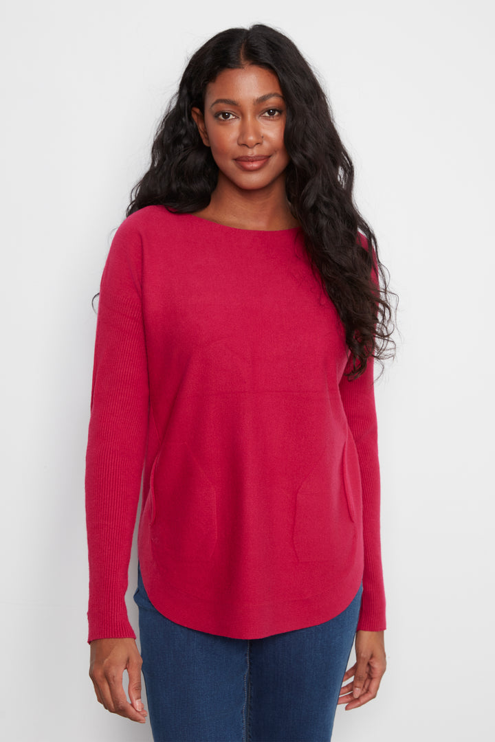 A woman modeling a bright pinkish-red long-sleeve knit sweater with a boat neckline and front patch pockets, paired with blue jeans against a plain light gray background.