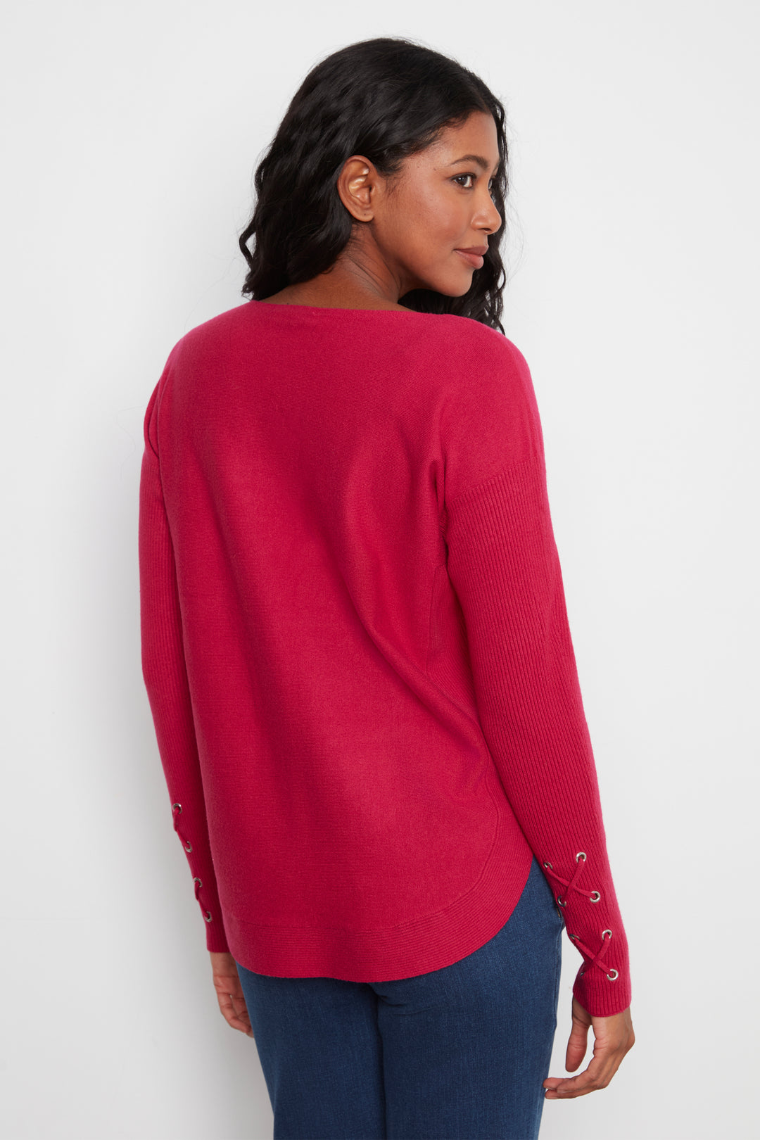 A woman wearing a bright pinkish-red long-sleeve knit sweater with lace-up details on the cuffs, shown from a three-quarters back view against a plain white background.