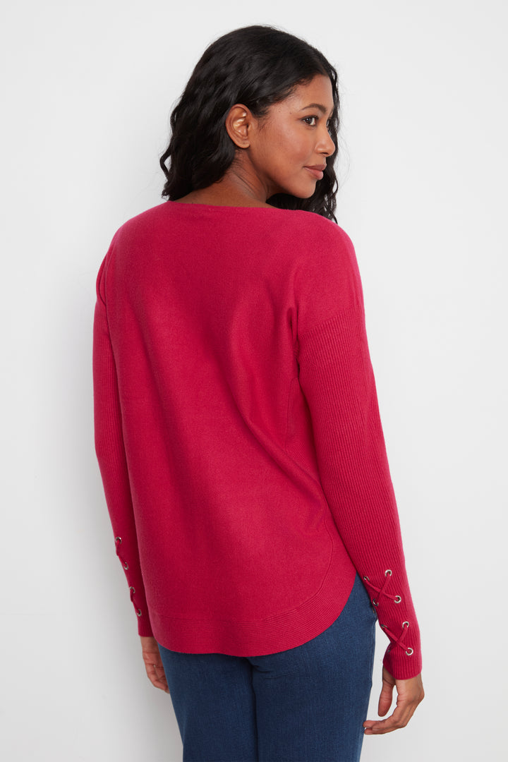 A woman wearing a bright pinkish-red long-sleeve knit sweater with lace-up details on the cuffs, shown from a three-quarters back view against a plain white background.