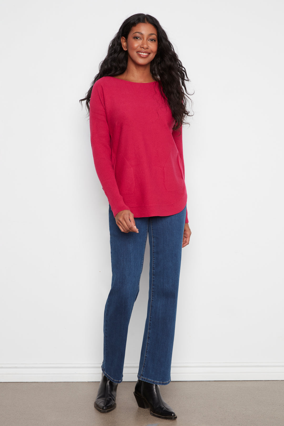 A woman modeling a bright pink long-sleeve knit top with a boat neckline, paired with blue jeans and black ankle boots, standing against a plain white wall.