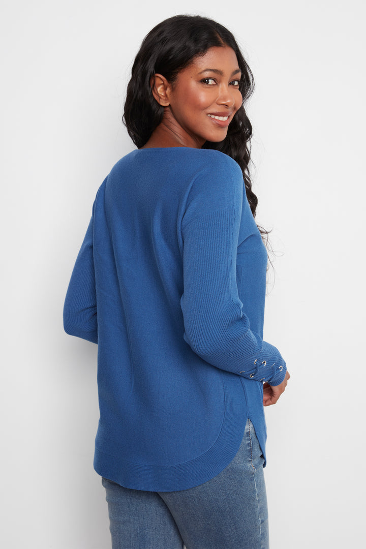 A woman wearing a blue ribbed knit long-sleeve top, shown from a three-quarter back angle against a plain white studio background.