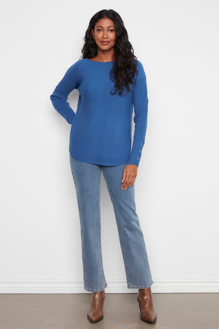 A woman modeling a bright blue long-sleeve knit top with a boat neckline, paired with light-wash blue jeans and brown ankle boots, standing against a plain white wall.
