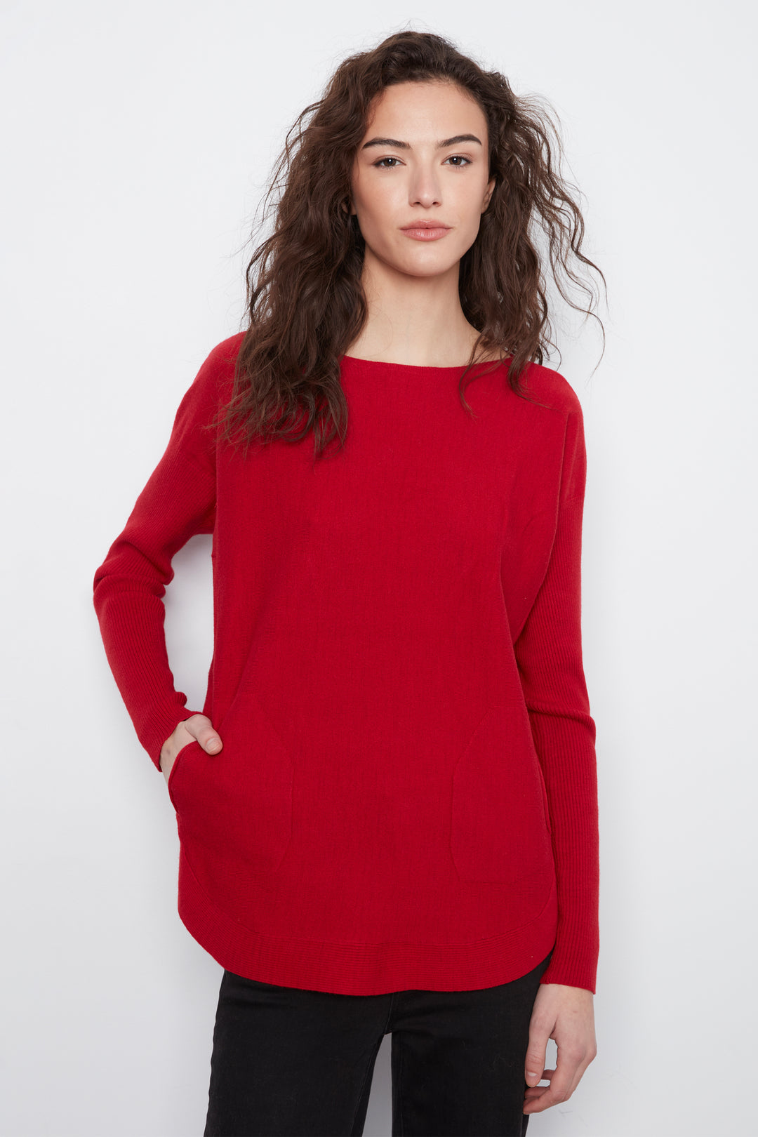 A woman modeling a bright red long-sleeve knit top with a boat neckline and side pockets, paired with black pants, against a plain light gray background.