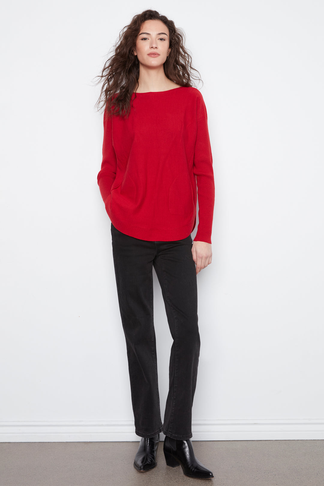 A woman modeling a bright red knit sweater with a wide boat neckline, paired with black trousers and black ankle boots, standing against a white wall.