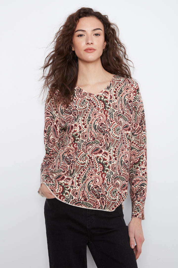 A woman modeling a long-sleeve V-neck blouse with an intricate paisley pattern in red, green, and beige, paired with black pants, against a plain white background.