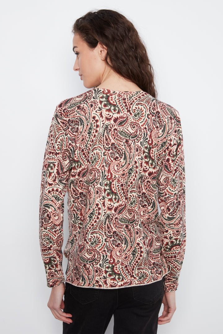 A woman viewed from the back wearing a long-sleeve top with an intricate paisley pattern in dusty pink, rust red, olive green, and cream on a light beige base, paired with black pants against a plain white background.