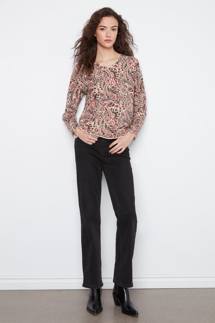 A woman stands against a white wall wearing a long-sleeve paisley blouse with black pants and black ankle boots.