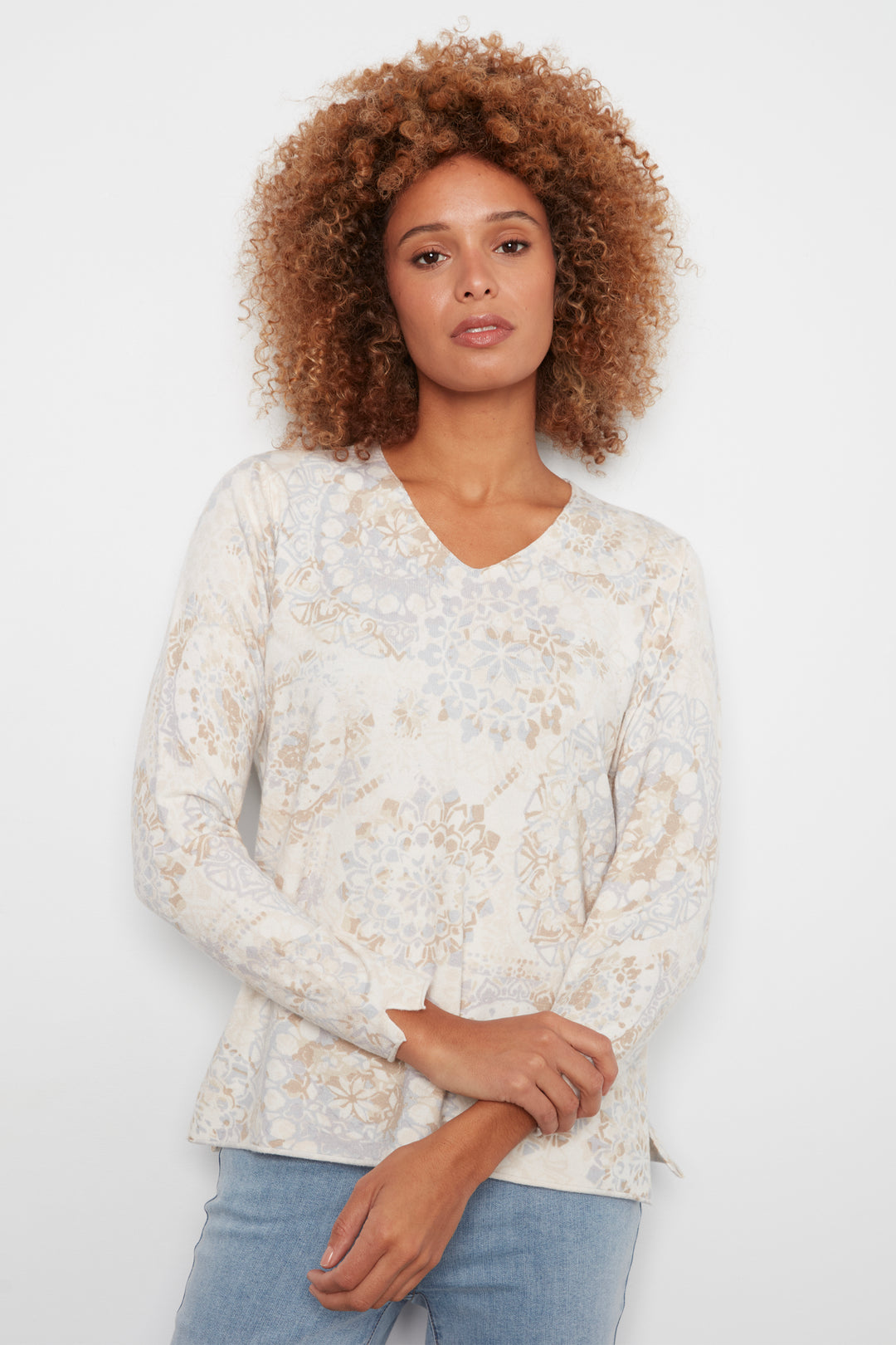 A woman with curly light brown hair wearing a cream-colored long-sleeve blouse with a beige and light blue floral pattern, paired with light blue jeans, against a plain white background.
