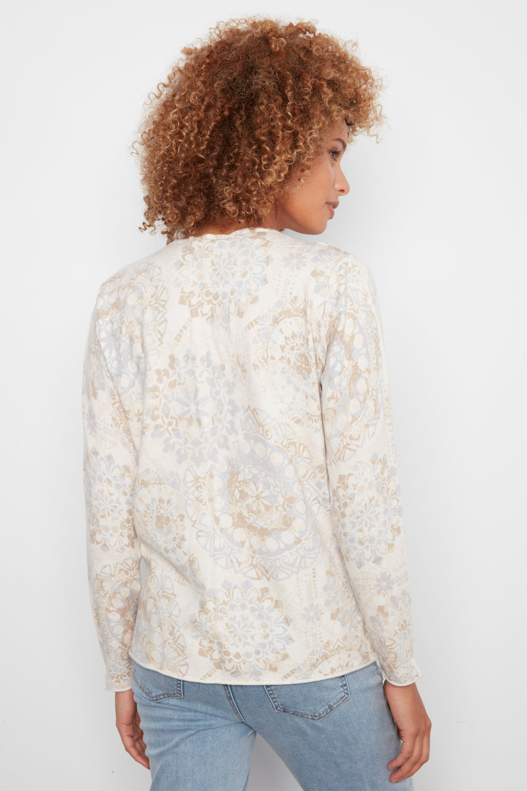 A woman with curly light brown hair is shown from the back wearing a cream-colored long-sleeve top with a beige and light blue paisley pattern, paired with light blue jeans, against a plain white background.