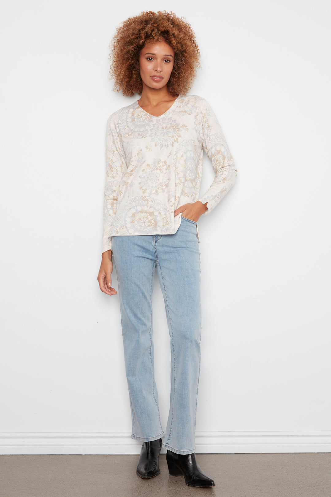 A woman modeling a cream-colored long-sleeve knit top with a delicate beige paisley pattern, paired with light-wash blue jeans and black ankle boots, standing against a white wall.