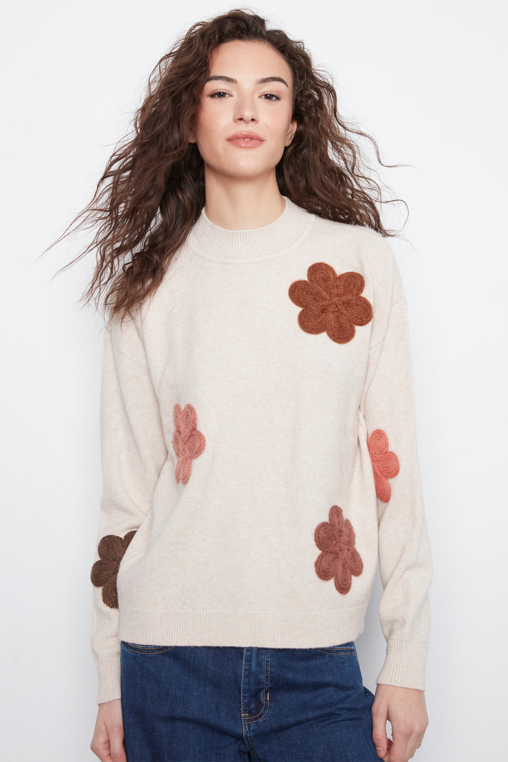A woman wearing a cream-colored knit sweater with brown and pink floral appliqués, paired with blue jeans, against a plain white background.