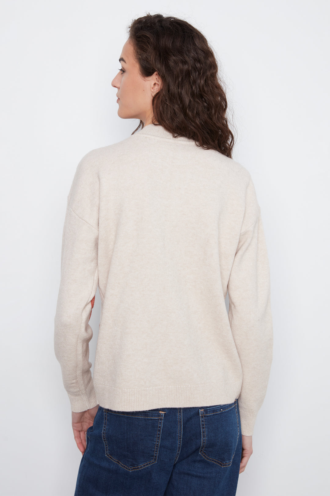 A woman with long, wavy brown hair is shown from the back wearing a light beige knit sweater and dark blue jeans against a plain white background.
