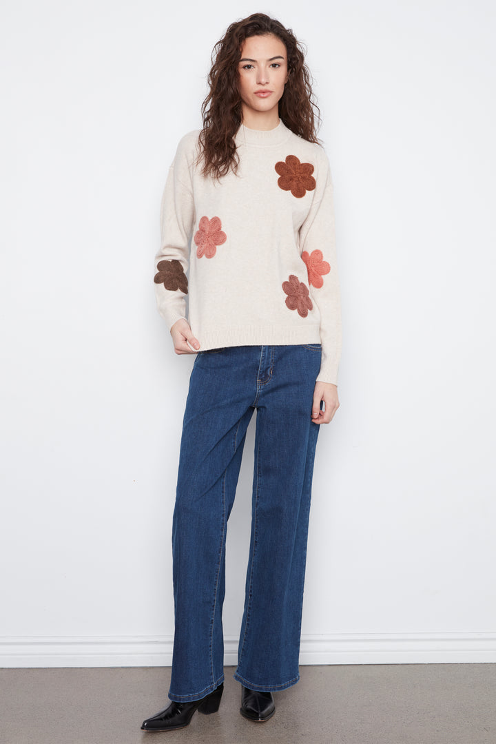 A woman stands against a white wall wearing a cream knit sweater with large brown and dusty rose flower appliqués, blue wide-leg jeans, and black pointed shoes.