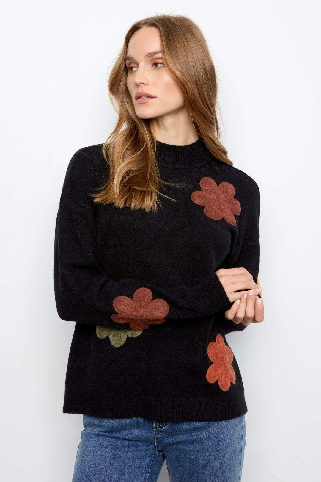 A woman wearing a black mock-neck knit sweater with brown and green floral appliqués, paired with blue jeans, photographed against a plain white studio background.