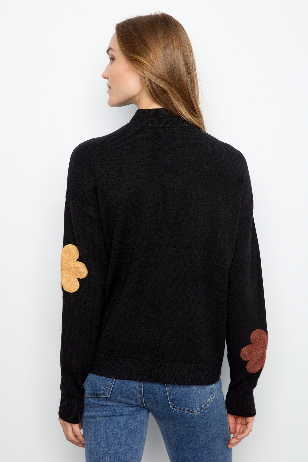 A woman shown from the back wearing a black knit sweater with tan and brown floral patchwork details on the sleeves, paired with blue jeans, against a plain white background.