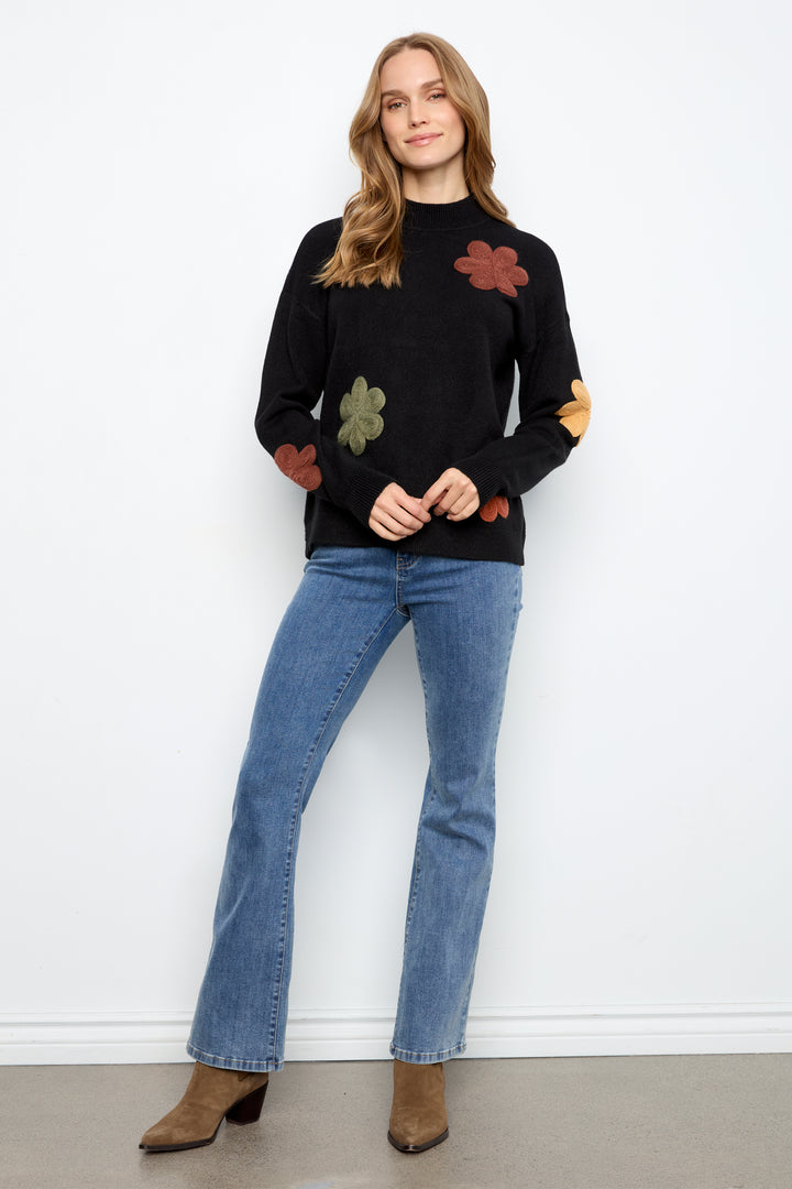 A woman modeling a black knit sweater with colorful embroidered flowers, paired with blue jeans and brown ankle boots, standing against a white wall.