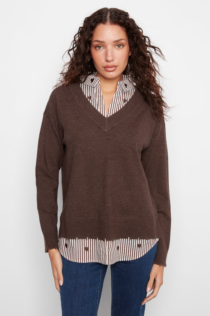 A woman modeling a brown V-neck knit sweater layered over a cream and brown striped shirt, paired with blue jeans, against a plain light gray background.