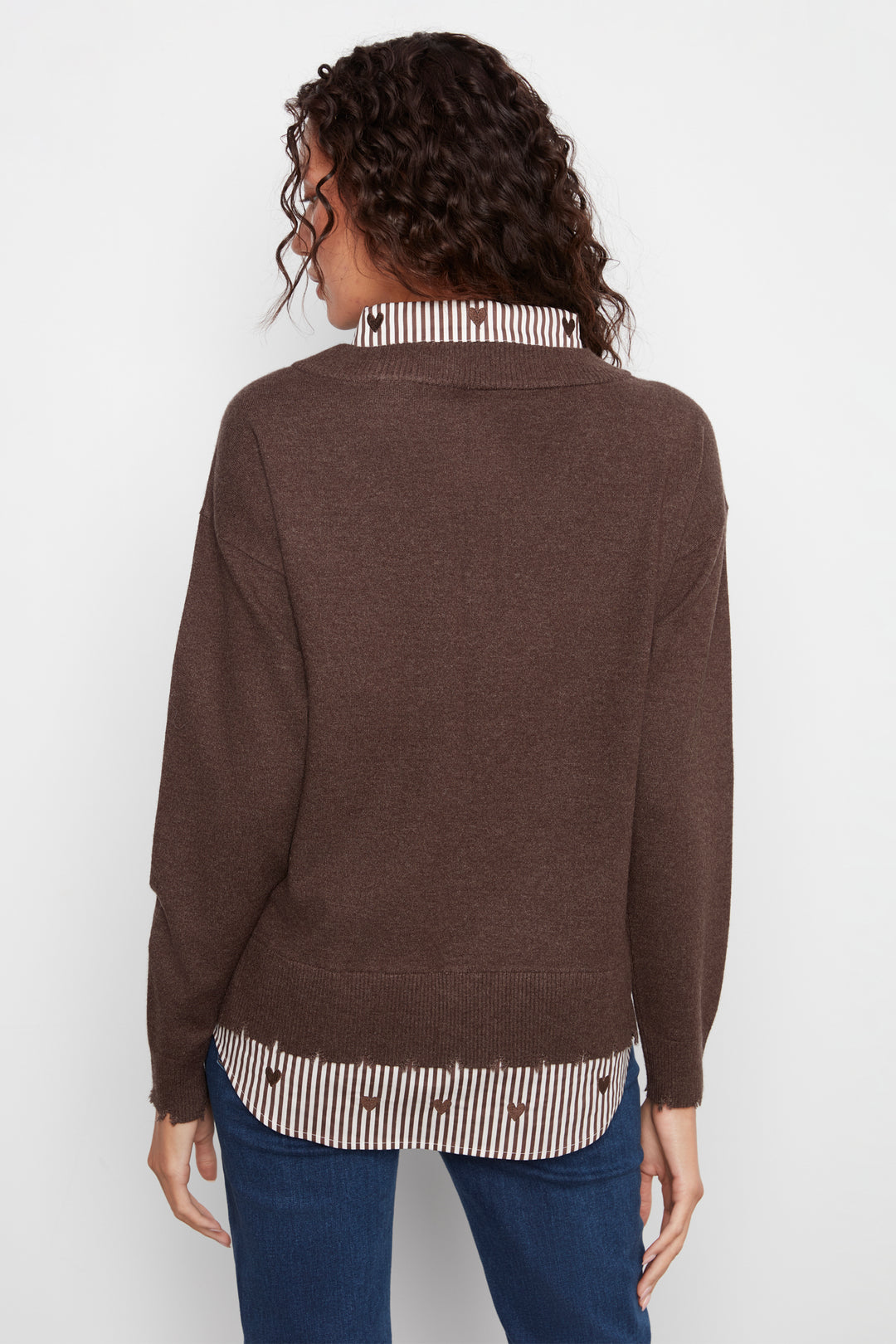 A woman shown from the back wearing a brown knit sweater over a striped shirt and blue jeans, photographed against a plain light background.