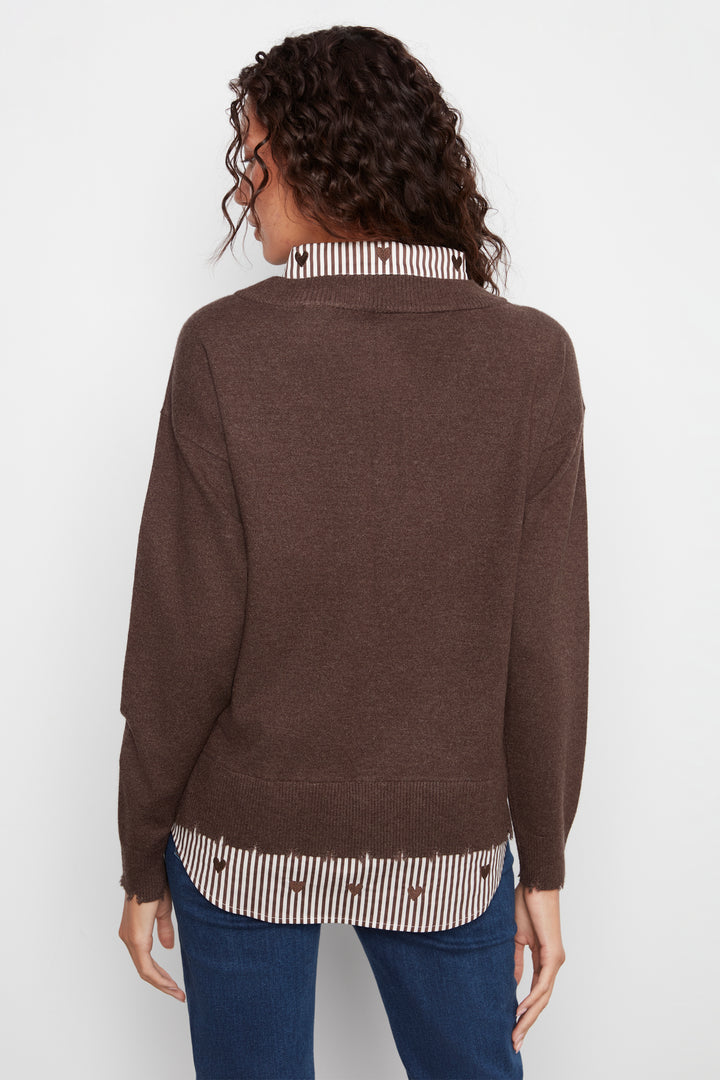 A woman shown from the back wearing a brown knit sweater over a striped shirt and blue jeans, photographed against a plain light background.