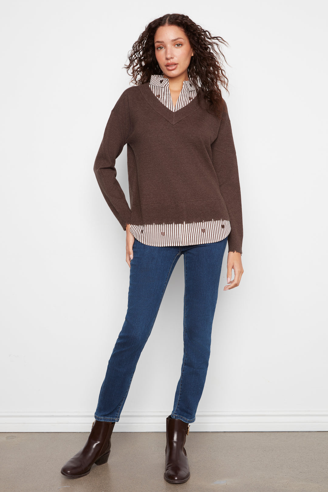 A woman modeling a brown v-neck knit sweater layered over a striped button-up shirt, paired with blue jeans and dark brown ankle boots, standing against a plain white wall.
