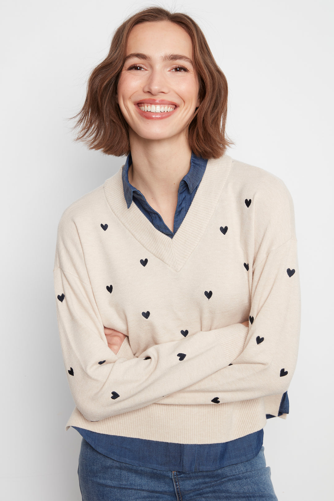 A smiling woman with short brown hair wearing a cream-colored V-neck sweater with small black heart patterns, layered over a blue collared shirt and blue jeans, against a light gray background.