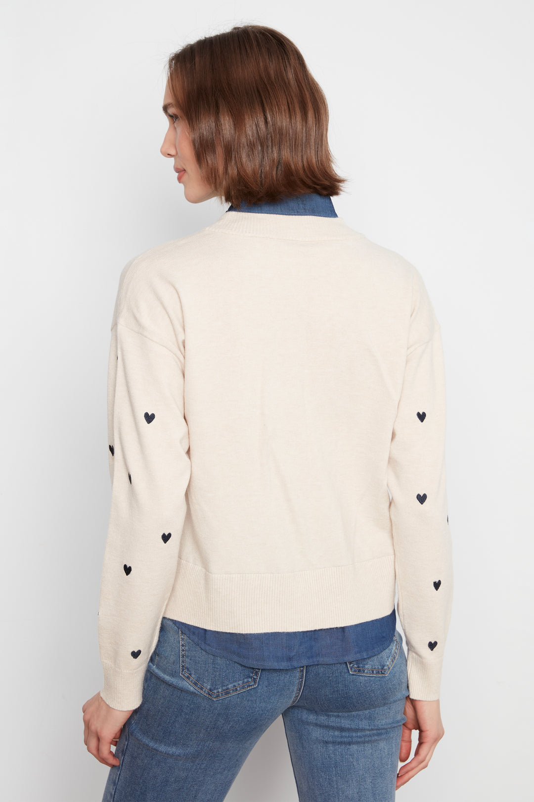 A woman shown from the back wearing a cream-colored knit sweater with small black heart details on the sleeves, layered over a blue collared shirt and paired with blue jeans, against a plain light gray background.