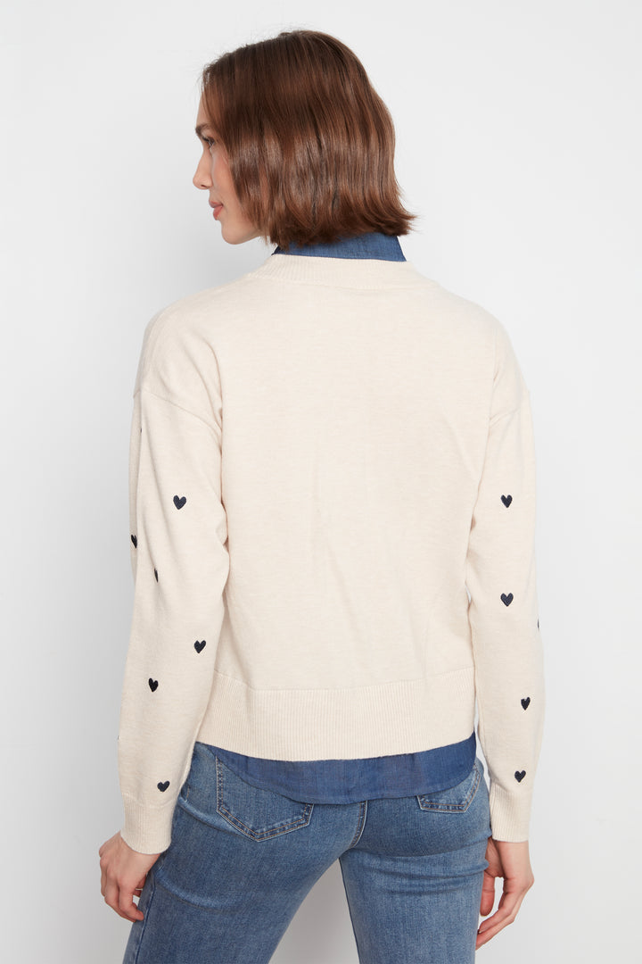 A woman shown from the back wearing a cream-colored knit sweater with small black heart details on the sleeves, layered over a blue collared shirt and paired with blue jeans, against a plain light gray background.