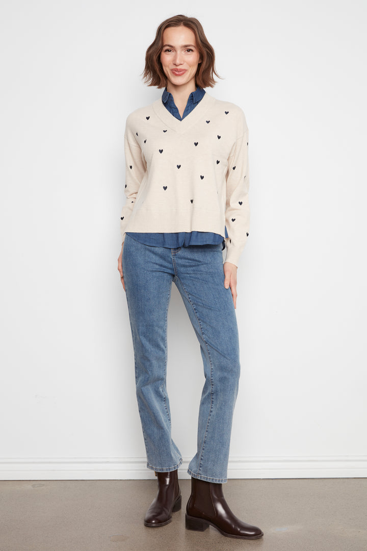 A woman stands against a white wall wearing a cream knit sweater with small black heart motifs, a blue collared shirt underneath, blue jeans, and dark brown ankle boots.