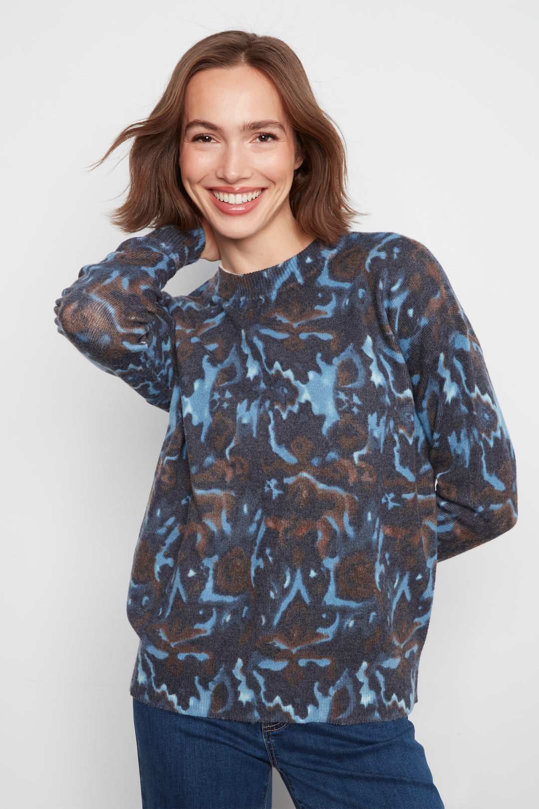 A woman wearing a long-sleeve crewneck sweater with an abstract blue, brown, and black pattern, paired with blue jeans, against a light gray studio background.