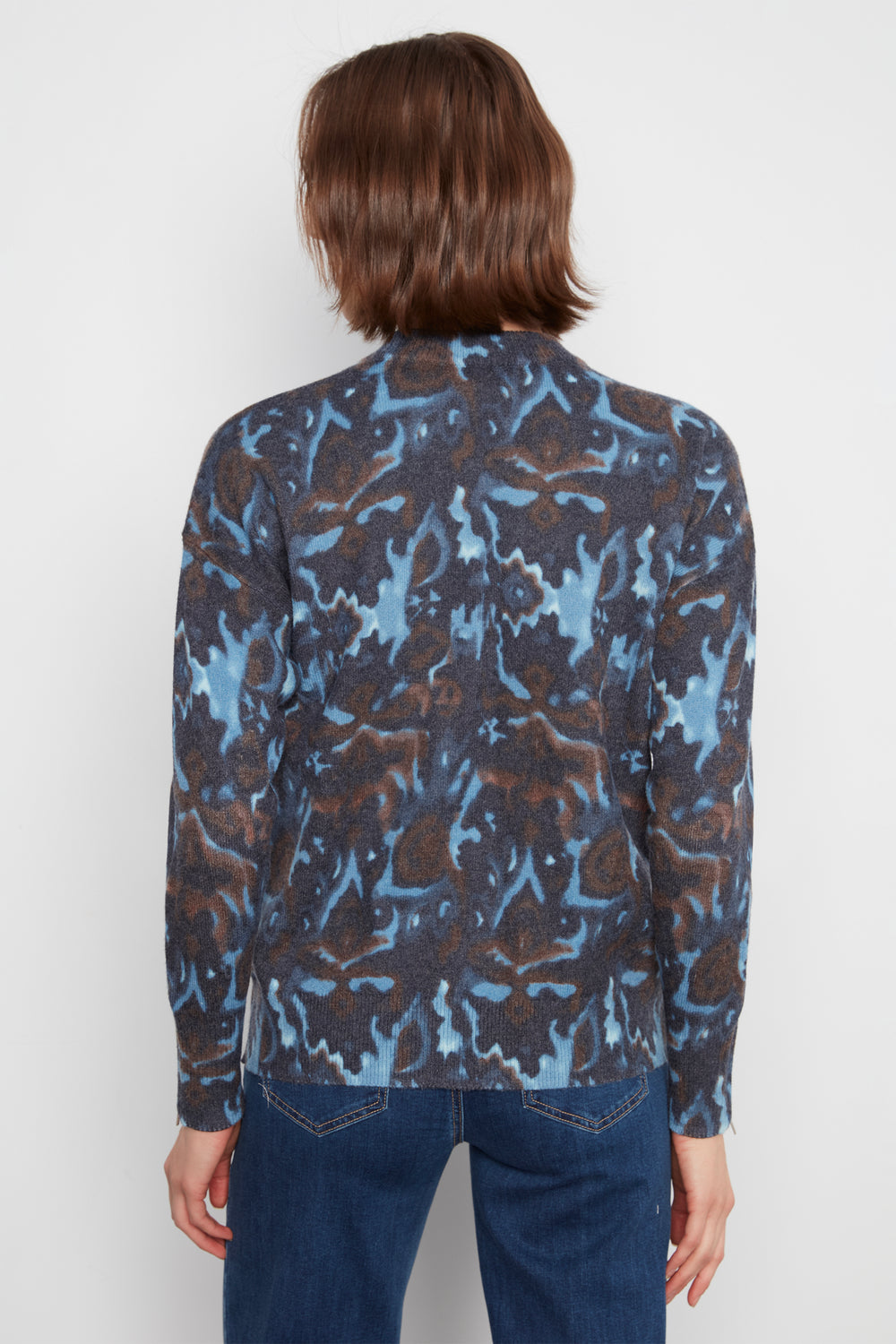 A person shown from the back wearing a long-sleeve button-front shirt with an abstract blue, brown, and black pattern, paired with dark blue jeans, against a light gray studio background.