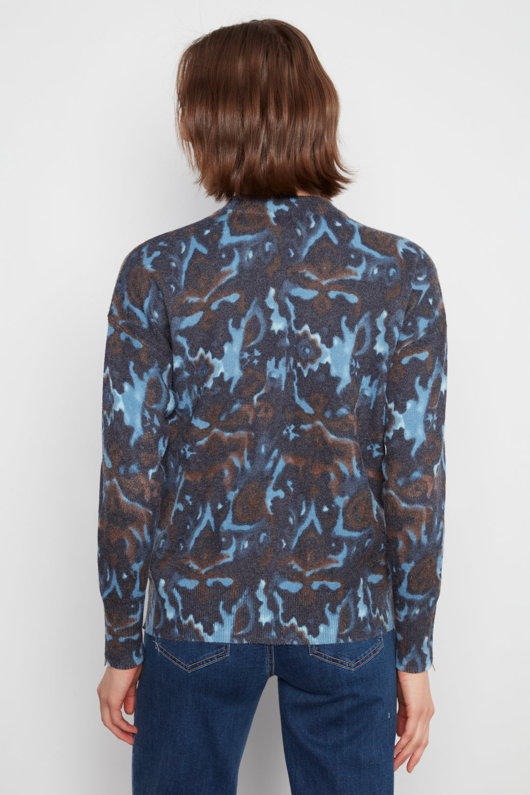 A person shown from the back wearing a long-sleeve button-front shirt with an abstract blue, brown, and black pattern, paired with dark blue jeans, against a light gray studio background.