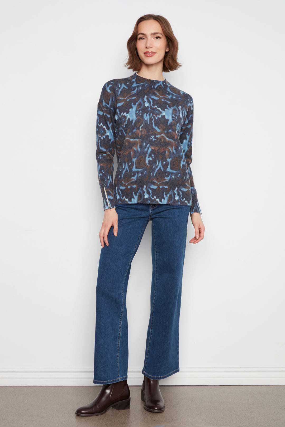 A woman stands against a plain white wall wearing a long-sleeve mock-neck blouse with a blue and brown abstract pattern, blue jeans, and dark brown leather boots.