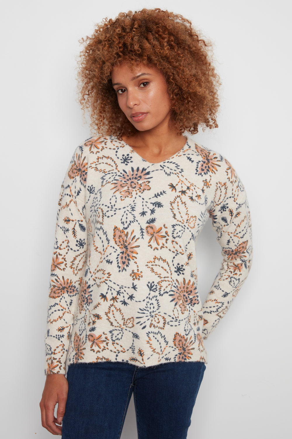 A woman with curly brown hair wearing a cream-colored long-sleeve knit top with a brown and navy floral pattern, paired with dark blue jeans, standing against a plain light gray background.