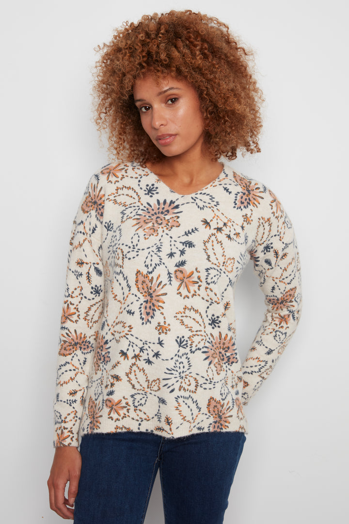 A woman with curly brown hair wearing a cream-colored long-sleeve knit top with a brown and navy floral pattern, paired with dark blue jeans, standing against a plain light gray background.