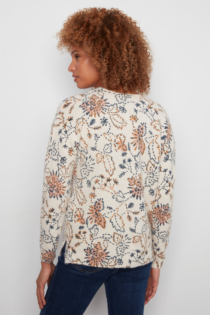 A woman with curly brown hair is shown from the back wearing a cream-colored knit sweater with a blue and rust-orange floral pattern, paired with dark blue jeans, against a plain light gray background.