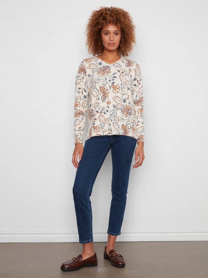 A woman stands against a white wall wearing a cream long-sleeve top with a brown and blue botanical pattern, dark blue skinny jeans, and brown leather loafers.