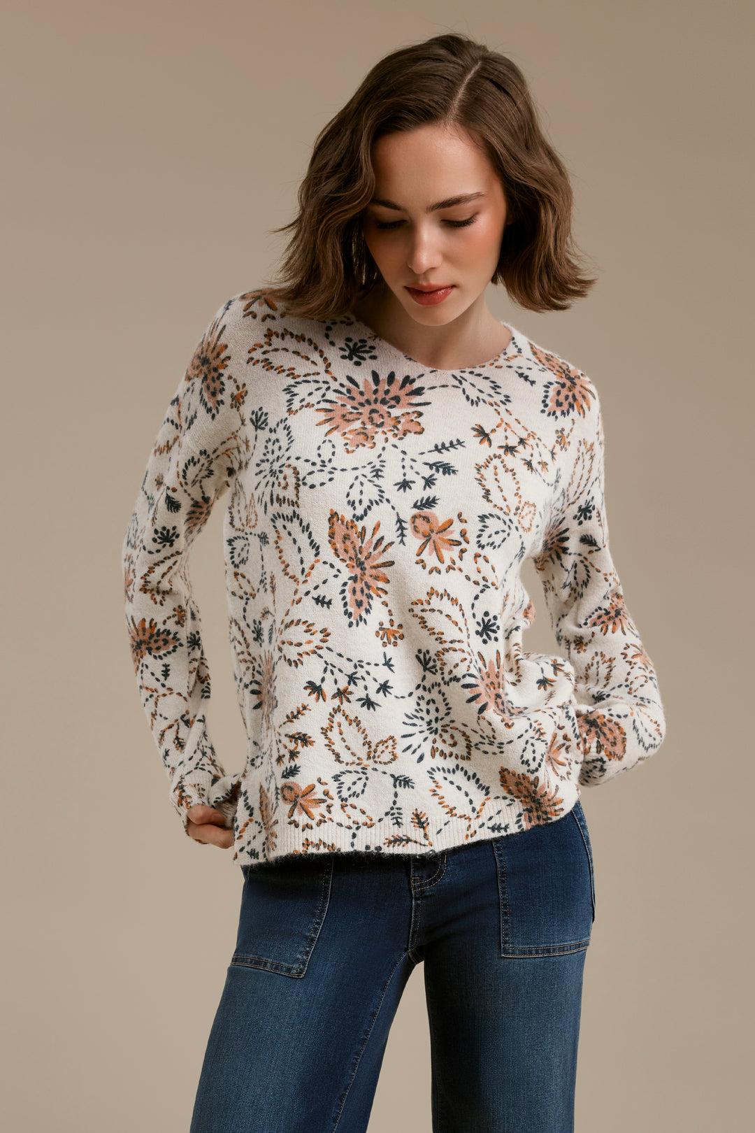A woman wearing a cream-colored knit sweater with a dense botanical and floral pattern in rust orange, navy blue, and black, paired with dark blue jeans, set against a solid warm beige studio background.
