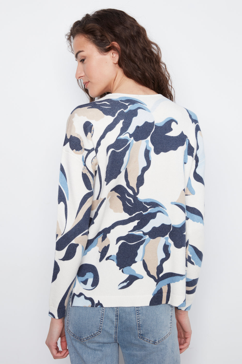 A woman shown from the back wearing a long-sleeve top with an abstract wavy pattern in navy blue, light blue, and beige on an off-white background, paired with light-wash blue jeans against a plain white studio background.