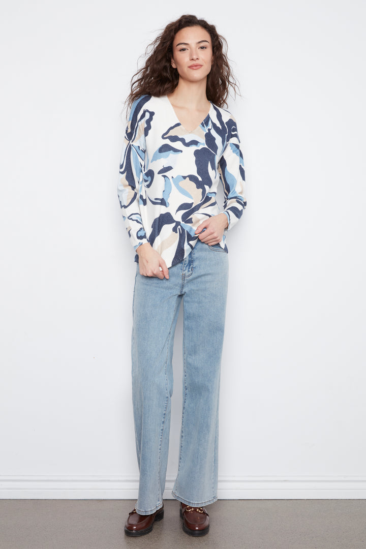 A woman stands against a white wall wearing a long-sleeve V-neck blouse with a blue, navy, and beige abstract floral pattern, paired with light-wash wide-leg jeans and dark brown loafers.