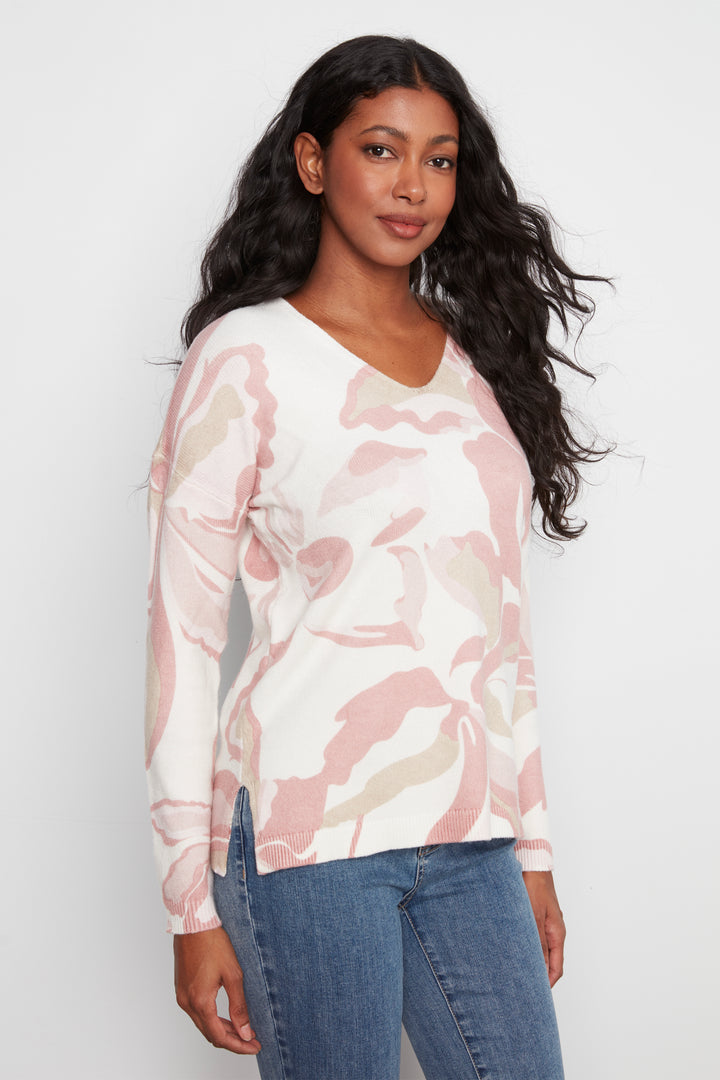 A woman modeling a long-sleeve V-neck knit top with a pink, beige, and white abstract floral pattern, paired with blue jeans, against a light gray studio background.
