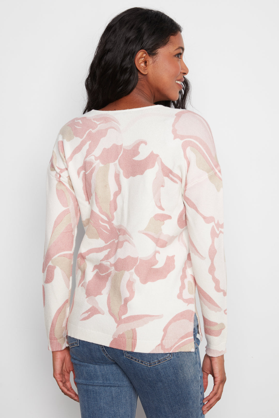 A woman shown from the back wearing a long-sleeve cream knit top with a pink and beige abstract floral pattern, paired with blue jeans, against a light gray studio background.